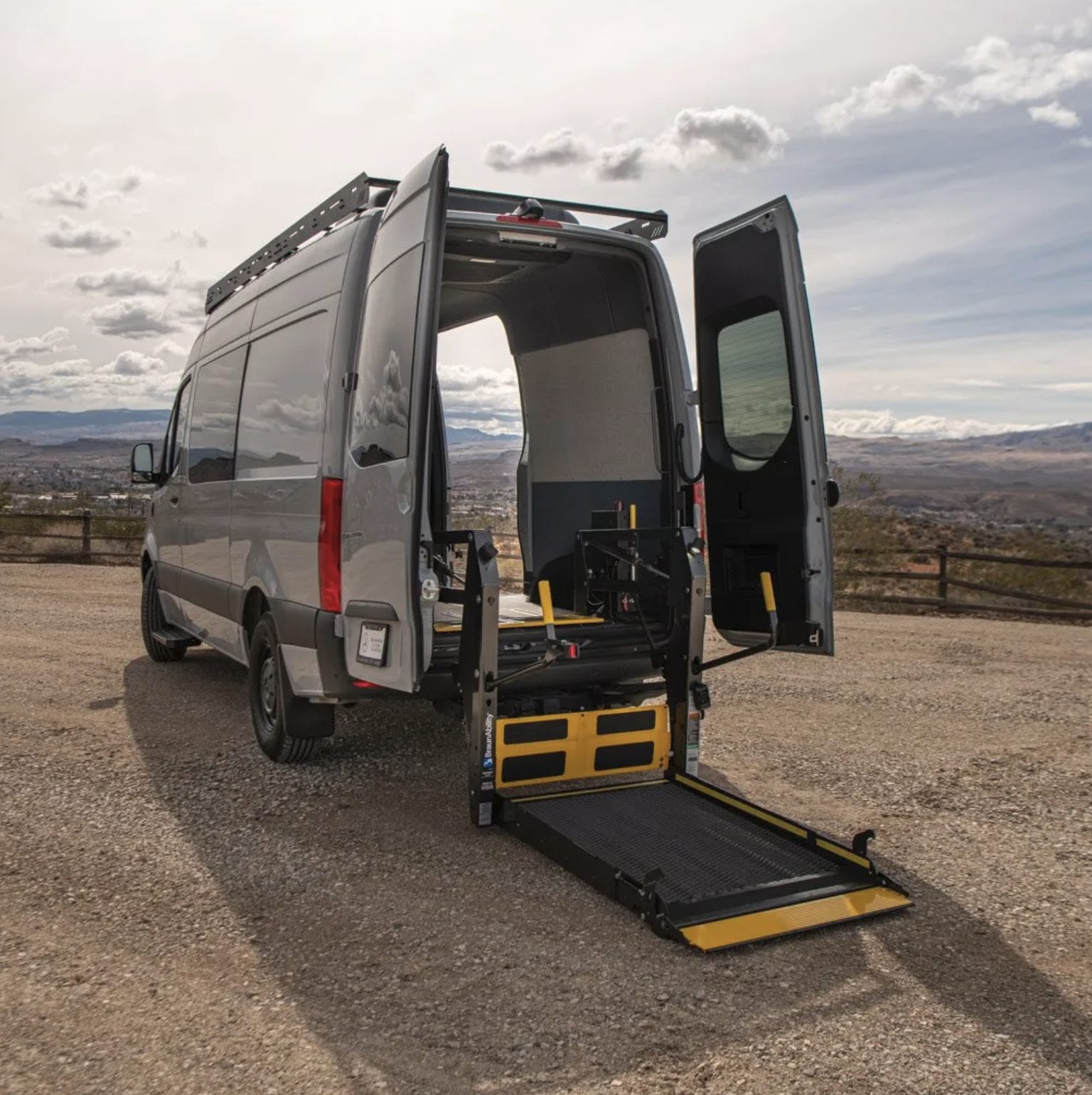 The in-stock silver Mercedes-Benz Sprinter photographed from the rear three-quarter on a gravel overlook, both rear doors open and the BraunAbility platform lift lowered flat to the ground.
